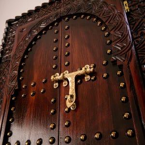 Puede incluir: Primer plano de una puerta de madera marrón oscuro con intrincadas tallas y detalles en latón. La puerta presenta un pestillo decorativo de latón y está tachonada con pequeños remaches redondos de latón. La veta de la madera es visible, añadiendo encanto rústico.