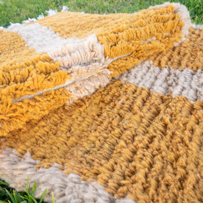May include: A close-up of a woven rug with a striped pattern in shades of beige and mustard yellow. The rug is made of thick, fluffy fibres and has a textured surface.
