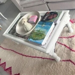May include: White coffee table with a glass top and a decorative metal design. The table has four legs and is sitting on a white and pink rug.