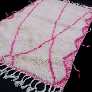 May include: White and pink Moroccan rug with a geometric pattern and fringe. The rug is made of wool and has a soft, fluffy texture.
