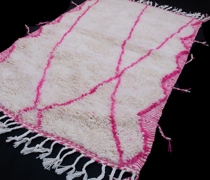 May include: White and pink Moroccan rug with a geometric pattern and fringe. The rug is made of wool and has a soft, fluffy texture.