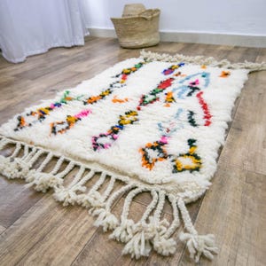 May include: A rectangular off-white wool rug featuring a high-pile texture and long, thick fringe on both ends. The surface is decorated with scattered, abstract geometric patterns in vibrant shades of orange, green, blue, pink, and yellow.