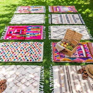May include: A collection of colorful, rectangular rugs with fringed edges are arranged on green grass. The rugs feature various geometric patterns and designs in shades of pink, white, blue, and brown. A picnic basket with fruit and a hat are also present.