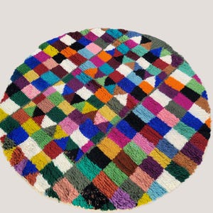 Könnte beinhalten: Ein runder, getufteter Teppich mit einem lebendigen, mehrfarbigen Schachbrettmuster. Das Design besteht aus kleinen, quadratischen Blöcken in verschiedenen Blau-, Grün-, Gelb-, Orange-, Rosa-, Lila-, Braun-, Schwarz- und Weißtönen, die eine dichte, strukturierte Oberfläche bilden.