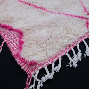 May include: White and pink Moroccan rug with a fluffy texture and fringe trim. The rug has a geometric pattern with pink stripes.