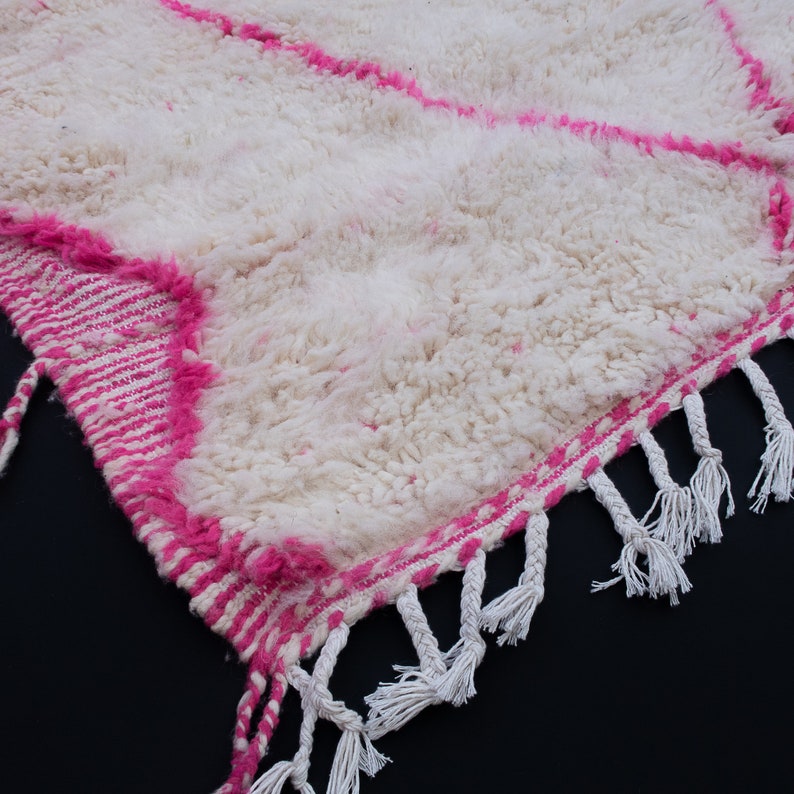 May include: White and pink Moroccan rug with a fluffy texture and fringe trim. The rug has a geometric pattern with pink stripes.