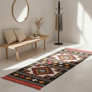 May include: A rectangular, patterned rug with a geometric design. The rug features a dark brown background with red, white, yellow, and black accents. The rug has a fringed edge and is placed on a light-colored floor.