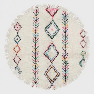Op de afbeelding: Een ronde, crèmekleurige vloerkleed met een gefranjerde rand. Het kleed heeft een geometrisch ontwerp met kleurrijke ruitvormen en verticale lijnen van kleine, kleurrijke bladvormige patronen. De diameter van het kleed is ongeveer 91 cm.