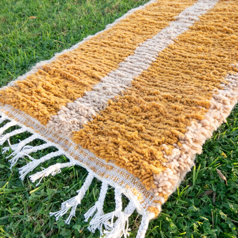 May include: A mustard yellow and cream striped rug with white fringe. The rug is made of a soft, fluffy material and has a hand-woven look.