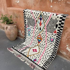 May include: A white rug with a geometric design featuring black, pink, and teal accents. The rug has a fringe border and is decorated with black dots, lines, and diamond shapes. The rug is leaning against a peach-colored wall.
