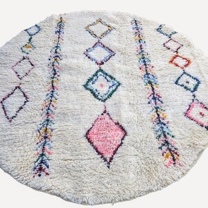 May include: A round, off-white area rug featuring a high-pile texture. The design includes two vertical, multi-coloured striped bands and a series of diamond-shaped motifs in various colours, including a prominent solid pink diamond in the centre.