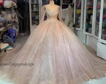 May include: A blush pink tulle ball gown with long sleeves and a beaded bodice. The gown has a sparkly finish and is shown on a dress form in a room with shelves of fabric.