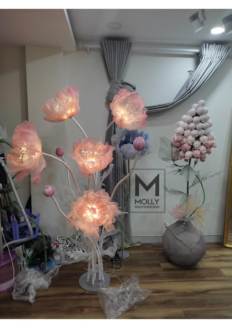 May include: A decorative floor lamp with pink fabric flowers and white stems. The lamp is lit with white lights and has a white base. The lamp is in a room with gray curtains and a white wall. The text "MOLLY NGUYENDESIGN" is visible on the wall.