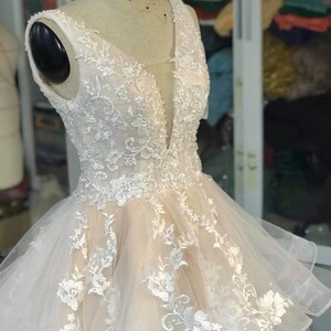 May include: A sleeveless, ivory wedding dress with a plunging V-neckline. The dress is made of a sheer fabric with delicate floral embroidery and a layered skirt.