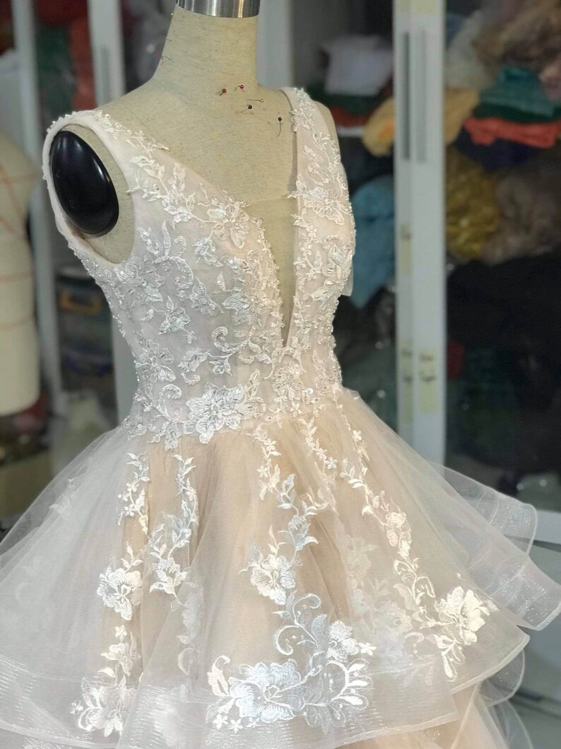 May include: A sleeveless, ivory wedding dress with a plunging V-neckline. The dress is made of a sheer fabric with delicate floral embroidery and a layered skirt.