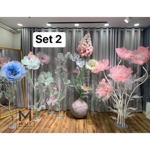 May include: Set of 2 large, decorative, fabric flowers. The flowers are in shades of pink, blue, and white. The flowers are on stands and are arranged in a room with gray curtains. The text "Set 2" is visible in the image.