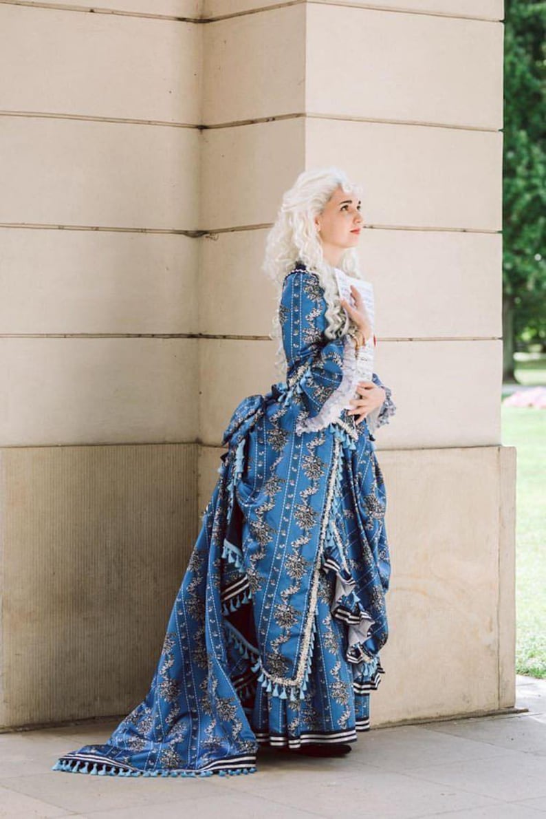 May include: A woman in a blue and white patterned dress with a long train and white lace trim. She is standing in front of a white wall and holding a piece of paper in her hand.