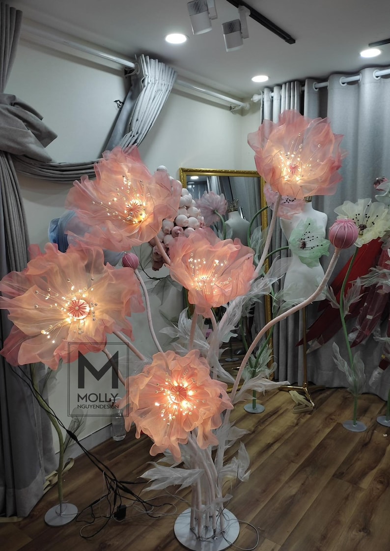 May include: A decorative light sculpture made of pink fabric flowers with white stems and lights. The flowers are arranged in a cluster and are lit from within. The sculpture is on a white base and is in a room with gray curtains.