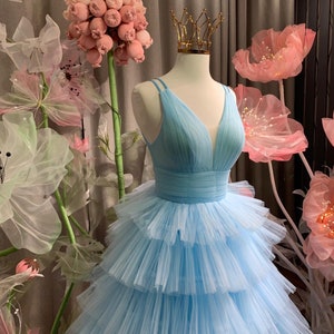May include: A light blue tulle dress with a V-neckline and a tiered skirt. The dress is on a mannequin and is surrounded by pink flowers.