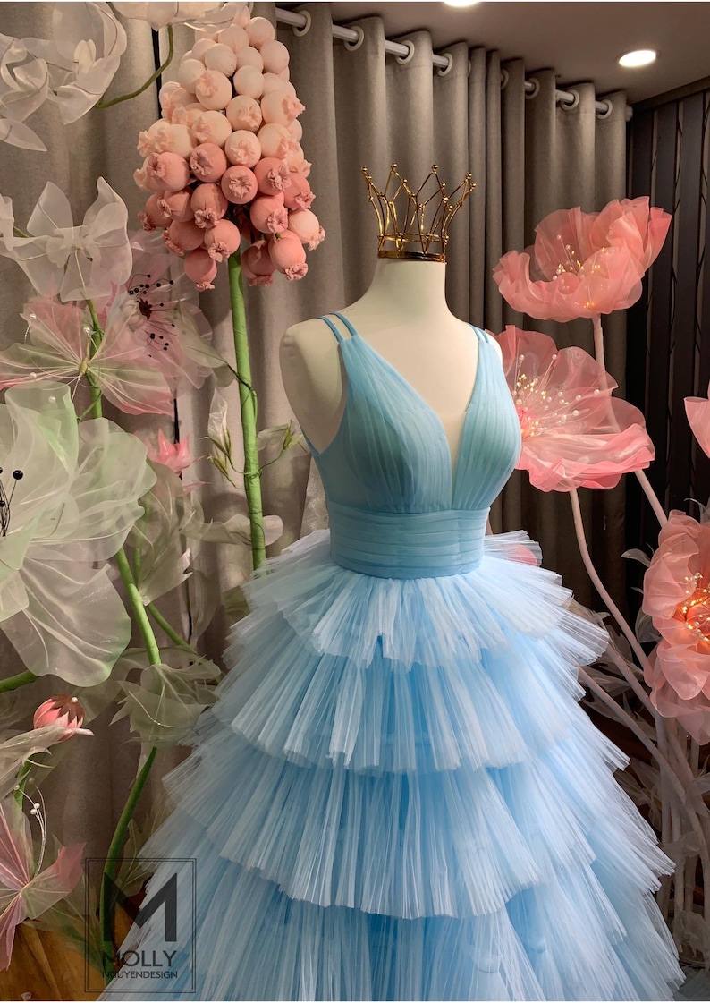 May include: A light blue tulle dress with a V-neckline and a tiered skirt. The dress is on a mannequin and is surrounded by pink flowers.