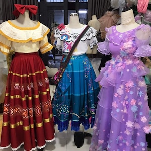 Puede incluir: Tres vestidos diferentes en maniquíes. El vestido de la izquierda es rojo con ribetes dorados y una blusa de encaje blanco. El vestido del medio es azul con una blusa blanca y un diseño de mariposa. El vestido de la derecha es morado con una blusa blanca y un diseño floral.