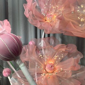 May include: Pink fabric flowers with white stems and pearl accents. The flowers have a light pink tulle fabric and are illuminated with warm white lights. The flowers are arranged in a cluster and are a decorative element for a wedding or event.