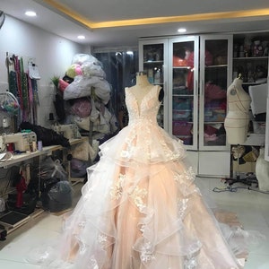 May include: A peach colored wedding dress with white lace detailing. The dress has a sweetheart neckline and a layered skirt. The dress is displayed on a mannequin in a room with a white tiled floor.