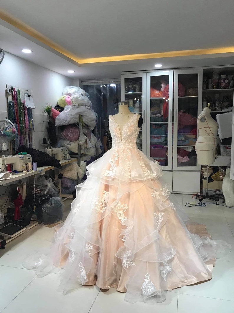 May include: A peach colored wedding dress with white lace detailing. The dress has a sweetheart neckline and a layered skirt. The dress is displayed on a mannequin in a room with a white tiled floor.