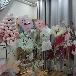 May include: Five large paper flowers on stands, in shades of pink, green, and yellow. The flowers are made of delicate, translucent fabric and have a whimsical, ethereal quality. The flowers are arranged in a room with gray curtains and a mirror.