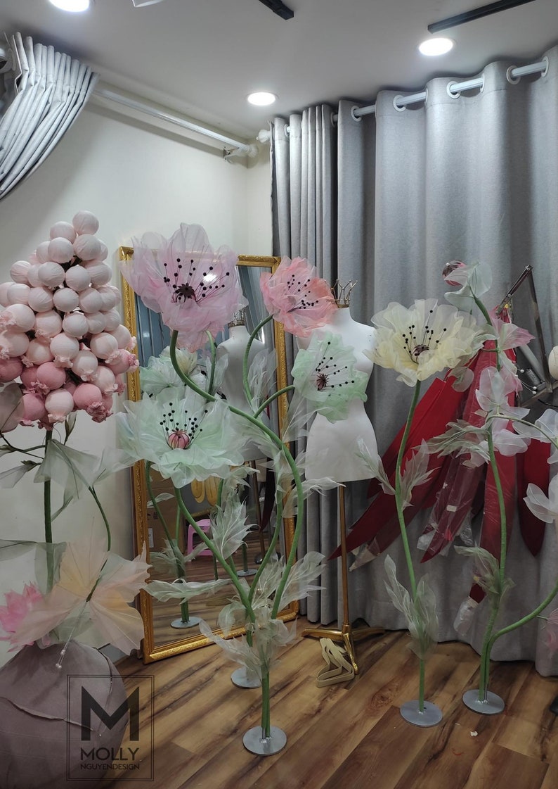 May include: Five large paper flowers on stands, in shades of pink, green, and yellow. The flowers are made of delicate, translucent fabric and have a whimsical, ethereal quality. The flowers are arranged in a room with gray curtains and a mirror.