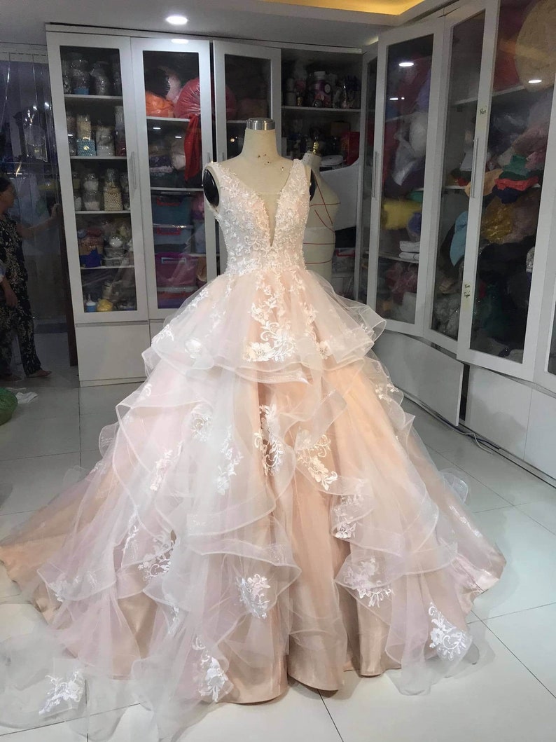 May include: A white and peach layered tulle wedding dress with delicate lace embroidery. The dress has a sweetheart neckline and a fitted bodice. The skirt is full and flowing with multiple layers of tulle.