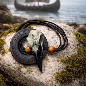 May include: A black raven skull pendant necklace with a circular, engraved pendant. The necklace features wooden beads and a black cord. A Viking ship sails in the background, set against a coastal landscape.