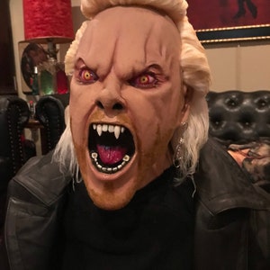May include: A close-up of a person wearing a costume mask of a vampire with a blonde wig, a black leather jacket, and a red tongue sticking out. The mask has red eyes and sharp teeth.