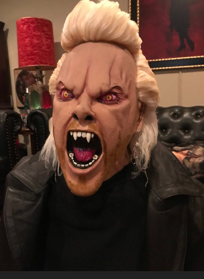 May include: A close-up of a person wearing a costume mask of a vampire with a blonde wig, a black leather jacket, and a red tongue sticking out. The mask has red eyes and sharp teeth.