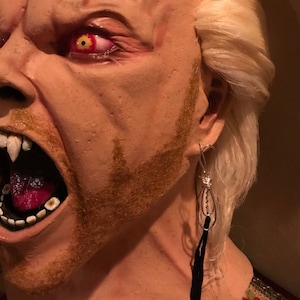 May include: A close-up of a plastic mask with a snarling expression, red eyes, and a fake beard. The mask is wearing a silver dangle earring with a black tassel.
