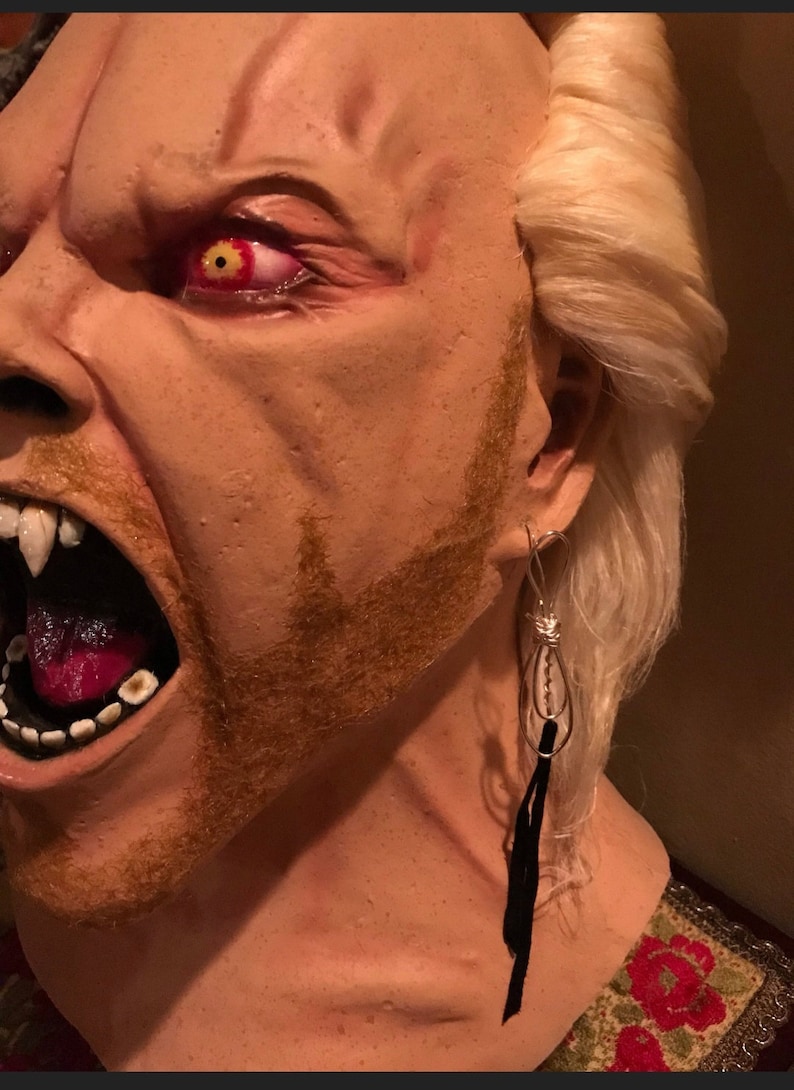 May include: A close-up of a plastic mask with a snarling expression, red eyes, and a fake beard. The mask is wearing a silver dangle earring with a black tassel.