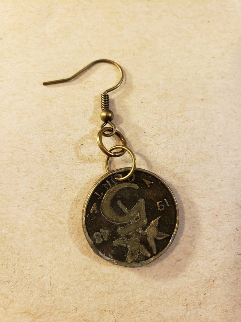 May include: A gold-toned earring featuring a round coin charm with a stylized letter 'G' and floral design. The coin charm has the text '1 LINO 51' around the edge.