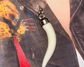 Puede incluir: Un pendiente individual que presenta un colgante liso y curvado en forma de colmillo de color blanco roto, suspendido de una tapa de metal plateado y un gancho estándar. El colgante tiene un acabado pulido y termina en una punta redondeada.