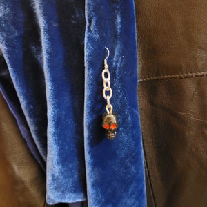 May include: A black skull earring with red eyes hanging from a silver chain. The earring is hanging from a blue velvet scarf.