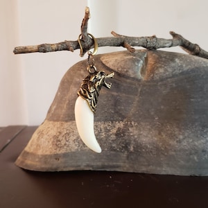 May include: A white tooth-shaped pendant with a gold wolf head charm. The pendant is hanging from a gold chain.