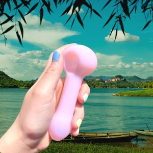 May include: A pink glass pipe with a rounded bowl and a long, slender stem. The pipe is held in a hand against a backdrop of a lake and mountains.