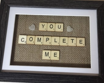 Könnte beinhalten: Gerahmtes Kunstwerk mit den Worten "YOU COMPLETE ME", die mit Scrabble-Steinen geschrieben wurden. Das Kunstwerk ist vor einem Jutegewebe-Hintergrund platziert, in einem dunkelgrauen Rahmen mit weißem Passepartout gerahmt und enthält zwei herzförmige Verzierungen.