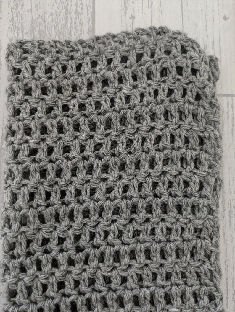 K&ouml;nnte beinhalten: Nahaufnahme eines grau gestrickten Stoffes mit offenem Gewebe. Der Stoff hat ein strukturiertes Aussehen mit einem gitterartigen Muster aus offenen R&auml;umen. Das Garn scheint eine mittelgraue Farbe zu haben.