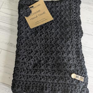 K&ouml;nnte beinhalten: Dunkelgraues, geh&auml;keltes Handtuch mit gewelltem Rand. Ein Etikett lautet "Crochet Hand Towel" und "100% Baumwolle Maschinenw&auml;sche". Ein kleiner Knopf und ein Etikett mit dem Wort "Mon" sind befestigt.