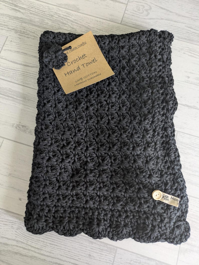 K&ouml;nnte beinhalten: Dunkelgraues, geh&auml;keltes Handtuch mit gewelltem Rand. Ein Etikett lautet "Crochet Hand Towel" und "100% Baumwolle Maschinenw&auml;sche". Ein kleiner Knopf und ein Etikett mit dem Wort "Mon" sind befestigt.