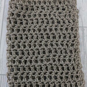 K&ouml;nnte beinhalten: Nahaufnahme eines handgestrickten Schals in neutralem Beige. Die offene Webart erzeugt ein gitterartiges Muster, das die Textur des Garns hervorhebt. Der Schal wirkt weich und flexibel, geeignet f&uuml;r verschiedene Stile.