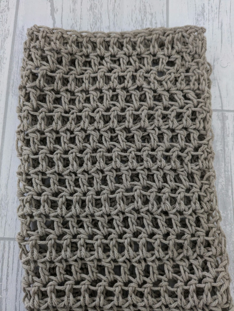 K&ouml;nnte beinhalten: Nahaufnahme eines handgestrickten Schals in neutralem Beige. Die offene Webart erzeugt ein gitterartiges Muster, das die Textur des Garns hervorhebt. Der Schal wirkt weich und flexibel, geeignet f&uuml;r verschiedene Stile.