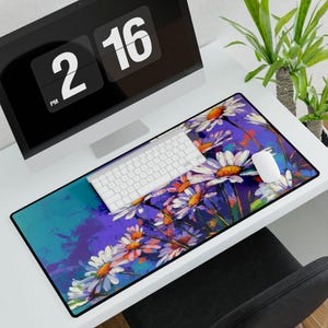 Pode incluir: Tapete de secretária retangular com um design floral de margaridas brancas com centros laranja, sobre fundos roxos e verde-azulados. Um teclado e um rato brancos estão sobre o tapete, com um monitor que mostra a hora 14:16.