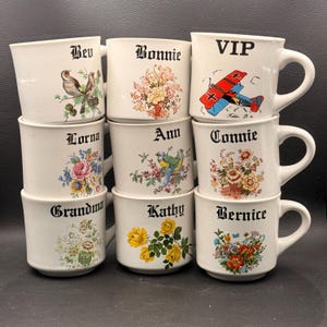 May include: A collection of white ceramic mugs with various designs and names. Each mug features a different floral or bird illustration, with names like "Bev," "Bonnie," and "VIP" in black gothic font. The mugs are stacked, showcasing their unique decorations.