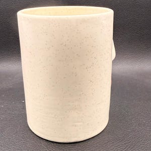 May include: A cylindrical ceramic mug with a speckled, cream-coloured finish. The mug has a smooth, rounded base and a straight, slightly flared top. The exterior is covered in small, dark speckles, giving it a rustic appearance. The mug is empty.